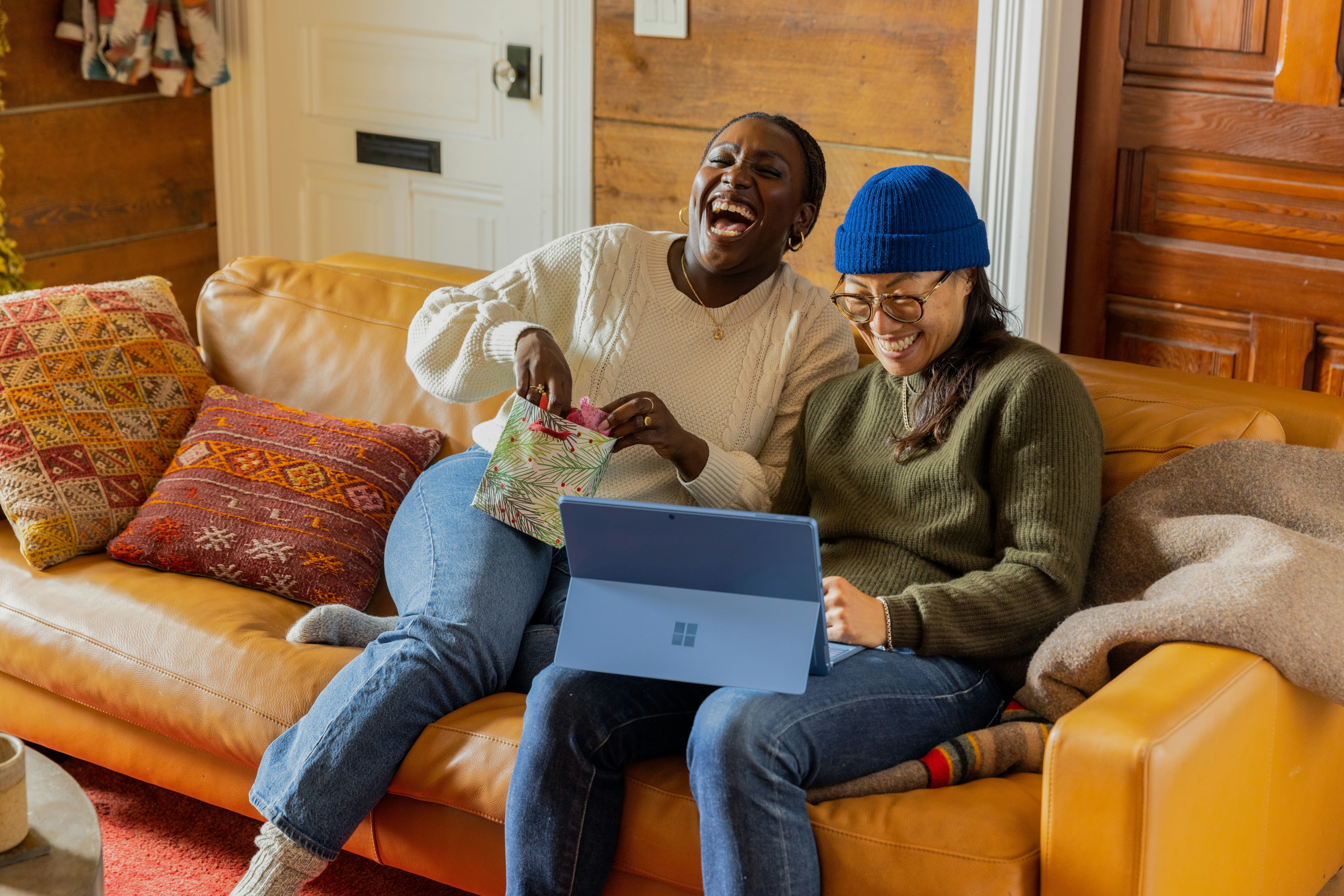 Duas mulheres sentadas no sofá sorrindo, uma está abrindo um presente e a outra utilizando o computador